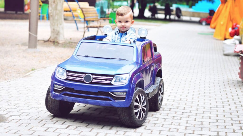 Malý chlapec řídí modré elektrické autíčko ve stylu terénního Jeepu na dlážděné cestě v parku.