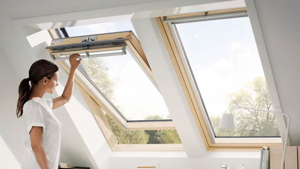Žena otevírá střešní okno Velux v moderním podkroví s výhledem do zeleně.